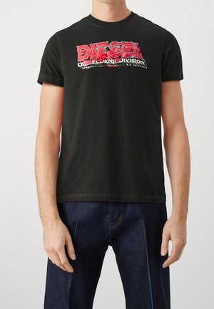 Mężczyzna ubrany w czarną koszulkę Diesel z czerwonobiałym logo w stylu distressed, w połączeniu z ciemnoniebieskimi dżinsami, stoi na jednolitym tle.