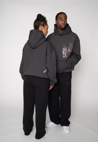 Deux personnes portent des sweats à capuche gris foncé assortis avec un graphisme de flammes et du texte. Ils sont assortis à des pantalons de survêtement noirs et des chaussures blanches.
