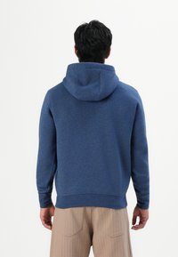 Man in een blauwe hooded sweatshirt en beige broek, staande met zijn rug naar de camera tegen een plain witte achtergrond.
