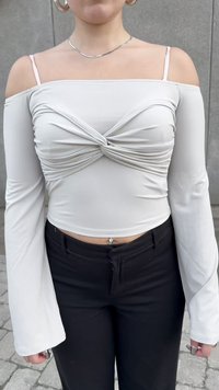 Mujer que lleva una blusa blanca de manga larga con los hombros al descubierto y un detalle de frente retorcido, pantalones negros, collar plateado, pendientes y anillos.