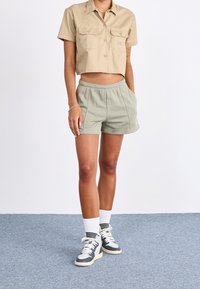 Béžová cropovaná košile na knoflíky se dvěma náprsními kapsami, v kombinaci s lehce zelenými sportovními šortkami a černo-b�ílými teniskami s bílými ponožkami.