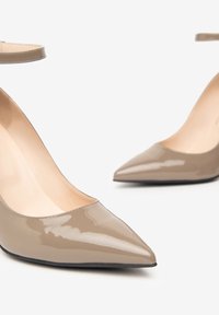 Lesklé béžové lodičky s špičatou špičkou a podpatkem, s kotníkovými pásky na bílém pozadí, ukazující detailní pohled na přední a boční stranu.