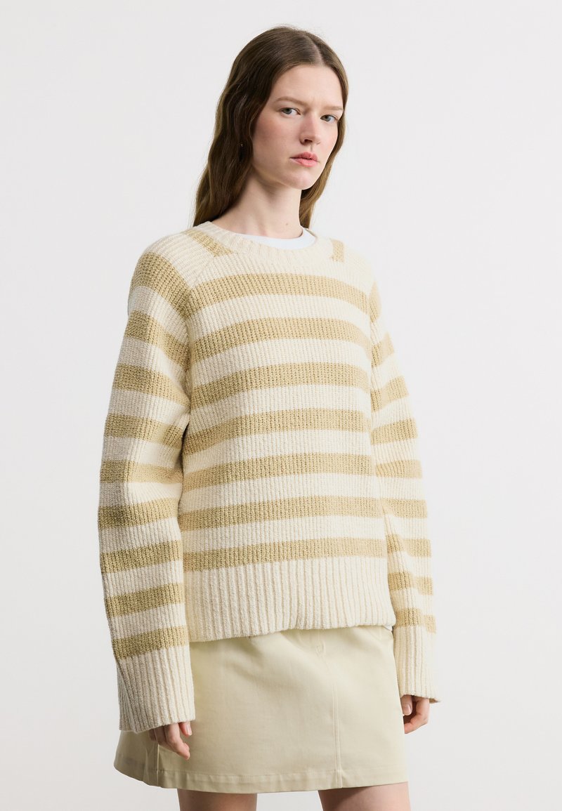 Γυναίκα που φοράει πουλόβερ πλεκτό με κρέμ και μπεζ ρίγες και μπεζ φούστα, στέκεται μπροστά σε ένα λευκό απλό φόντο.