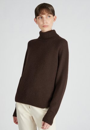 Maglione oversized marrone con collo a lupetto in tessuto morbido e testurizzato, con maniche lunghe e polsini a costine, abbinato a pantaloni chiari.