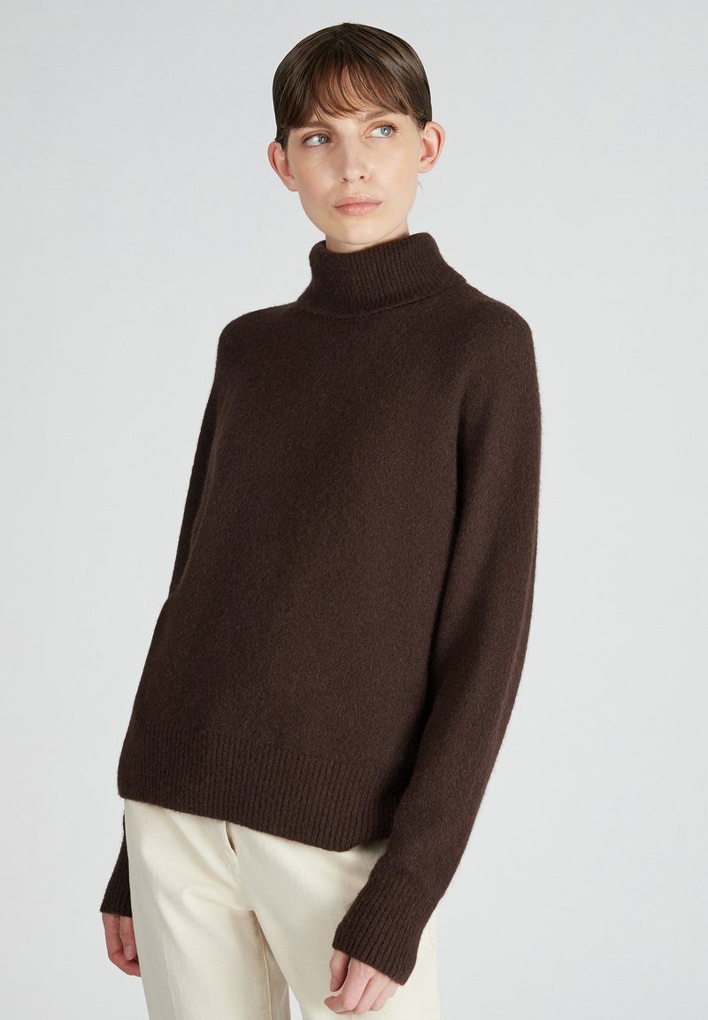 Maglione oversized marrone con collo a lupetto in tessuto morbido e testurizzato, con maniche lunghe e polsini a costine, abbinato a pantaloni chiari.