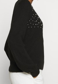 Svart långärmad tröja med pärldekorationer på bröstet, bärs med vita byxor, sidovy av person med handen i fickan.