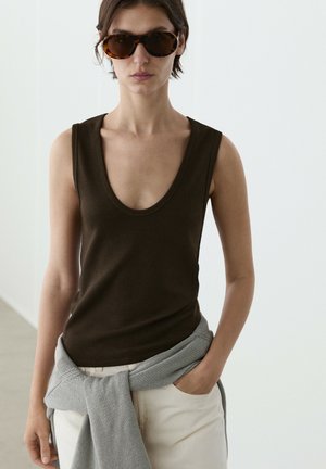 Jeune femme portant un top sans manches marron foncé, un pantalon beige, un pull gris noué autour de la taille, et des lunettes de soleil écaille, debout à l’intérieur.