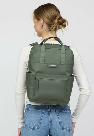 Frau mit blondem Haar zu einem Dutt gebunden, trägt ein weißes langärmliges Hemd und blaue Jeans, sie trägt einen grünen rechteckigen Rucksack mit Fronttasche.