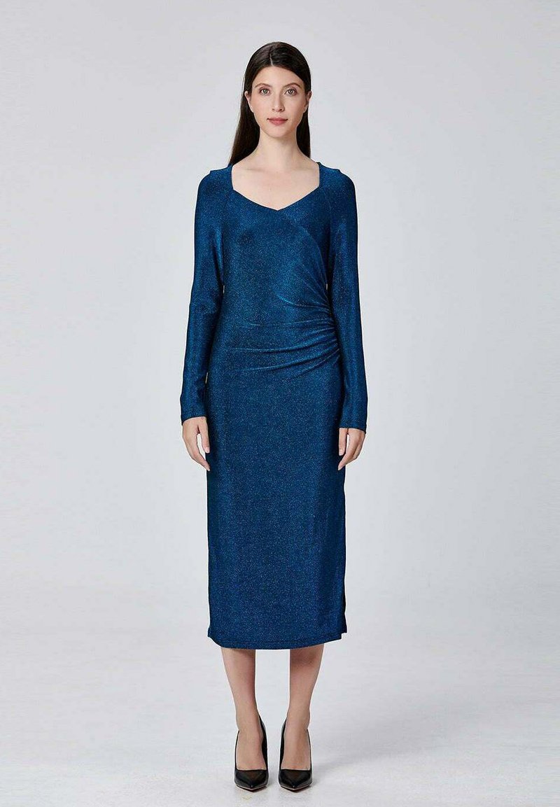 Femme se tenant droite, portant une robe brillante bleu foncé à manches longues et à hauteur des genoux, ainsi que des talons noirs, sur un fond clair uni.