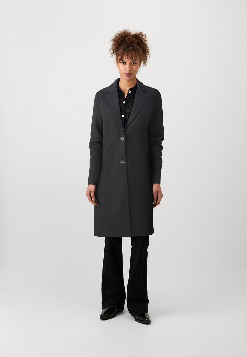 Long manteau noir avec une texture lisse, revers crantés, boutons avant, et une coupe ajustée, associé à un pantalon noir évasé et des chaussures brillantes.