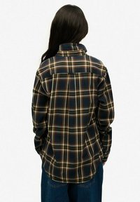 Chemise à carreaux bleue avec un col, des manches longues et une coupe ample. Le motif comprend des lignes jaunes, rouges et blanches sur un fond sombre.