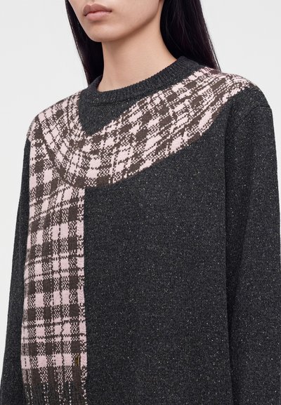 Femme portant un pull en maille gris foncé avec un motif à carreaux rose et brun sur l'épaule et le long d'un côté.