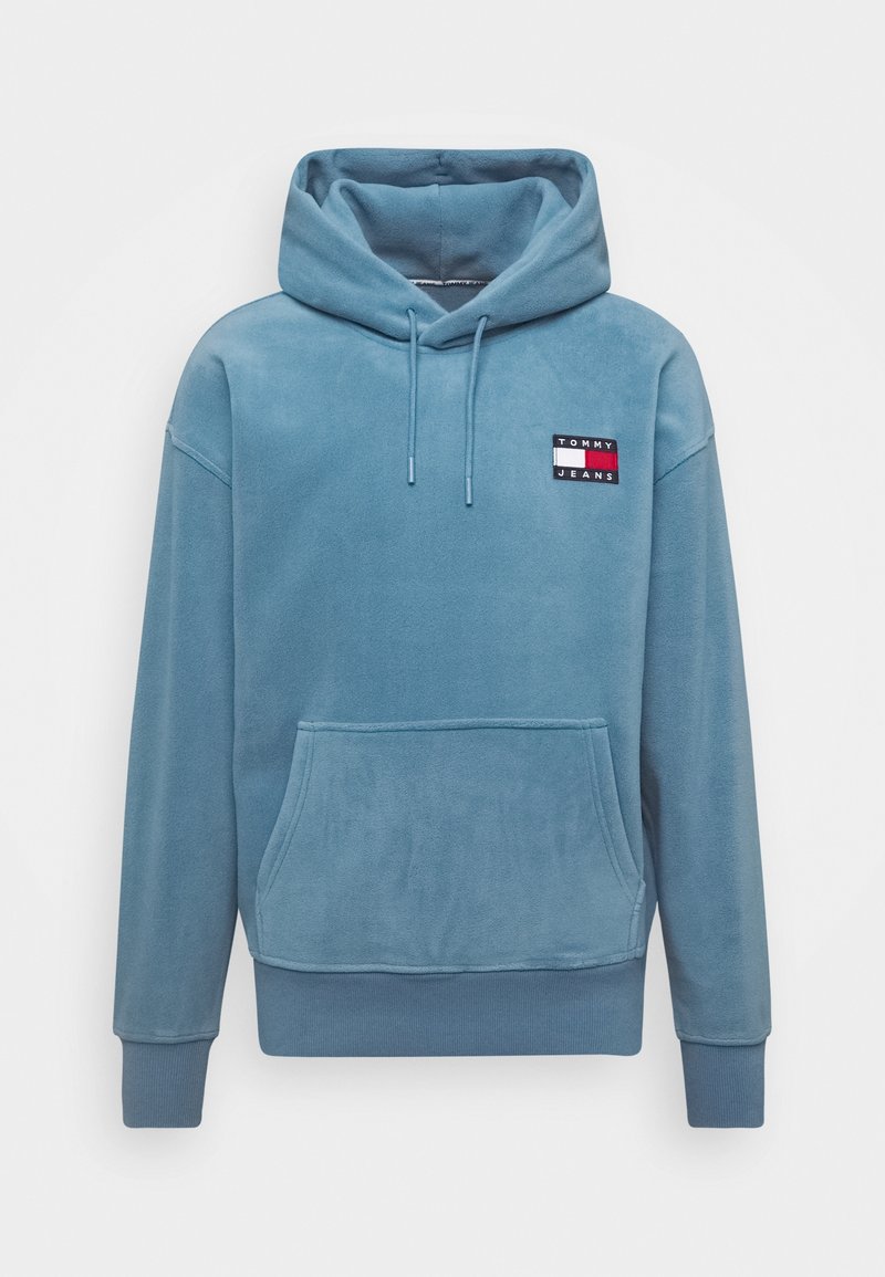 Sweat à capuche en polaire bleue avec une poche avant, des cordons et un petit écusson avec le logo "TOMMY JEANS" en rouge, blanc et noir sur la poitrine.