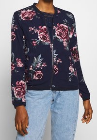 Marinblå blommig bomberjacka med rosa rosor, dragkedja framtill, ribbade cuffar och underkant, mjukt tyg, parat med ljusblå denimjeans.