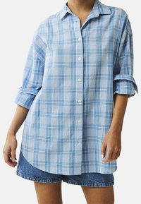 Světle modrá kostkovaná košile na knoflíky v uvolněném střihu, s límečkem a ohrnutými rukávy, spárovaná s džínovými šortkami.