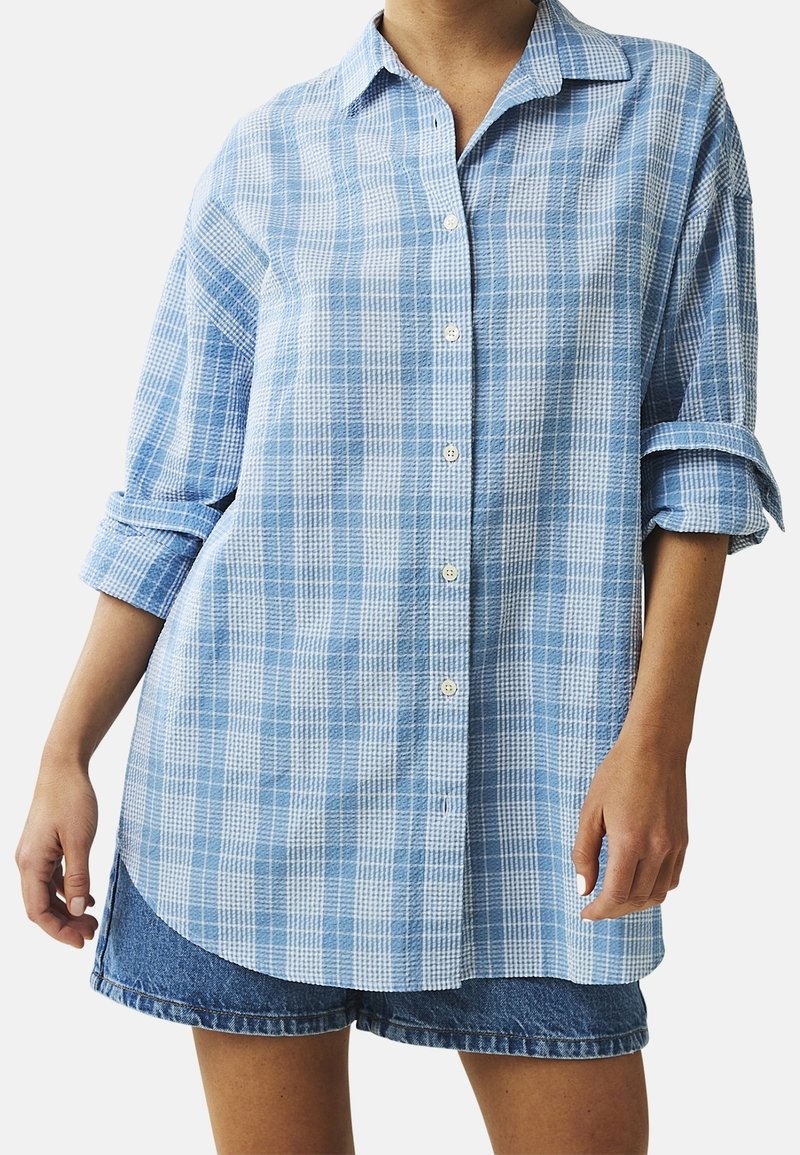 Světle modrá kostkovaná košile na knoflíky v uvolněném střihu, s límečkem a ohrnutými rukávy, spárovaná s džínovými šortkami.
