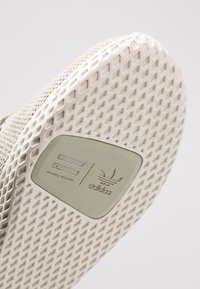 Sole boty Adidas ve světle béžové barvě, s texturovaným vzorem pro lepší přilnavost, vyrobená z gumového materiálu a s logem "Pharrell Williams" vytištěným nahoře.