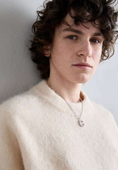 Κρέμας χρώματος, φουσκωτό πουλόβερ με στρογγυλή λαιμόκοψη. Διαθέτει ασημένιο κολιέ με στρογγυλό φυλαχτό στολισμένο με μικρά πετράδια.