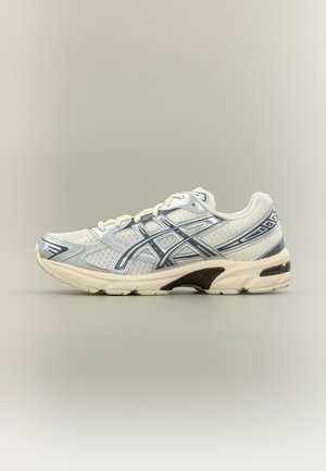 Biało-srebrny sportowy trampek z siateczkową cholewką, czarnymi akcentami na podeszwie i krzyżującymi się bocznymi paskami, pokazany w widoku bocznym na neutralnym tle.