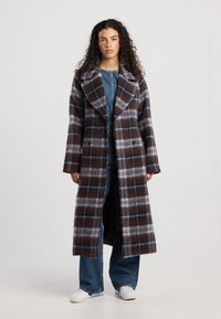 Brown and blue checked long coat with wide lapels and double-breasted buttons; soft, textured fabric; worn over denim and white sneakers.
