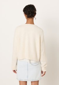 Krémově zbarvený svetr s texturou, pletený, s krátkým lemem a žebrovanými manžetami, doplněný světle modrou denimovou minisukní. Zobrazen pohled zezadu.