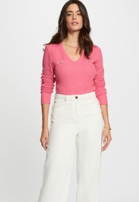 Femme avec de longs cheveux noirs portant un pull rose �à col V avec des boutons dorés et un pantalon large taille haute blanc, sur fond blanc.