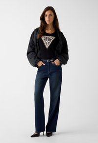 Czarna kurtka typu bomber na czarnym topie z trójkątnym logo "GUESS" wyszywanym cekinami, zestawiona z szerokimi niebieskimi dżinsami o kroju typu wide-leg i czarnymi szpilkami na szpic.