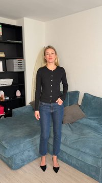 Moteris juodame kardigane ir mėlynuose džinsuose stovi svetainėje prie žalsvojo sofos ir šalia esančios juodos lentynos.