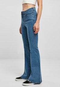 Jeans acampanados de color azul claro con cintura alta, que cuentan con un cierre de botón y cremallera, cinco bolsillos y costuras detalladas. Combinados con zapatillas negras.