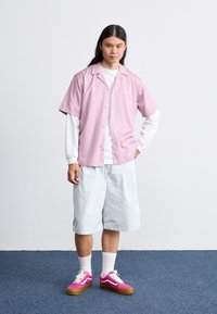 Chemise rose clair à manches courtes, t-shirt blanc à manches longues en dessous, shorts bleu clair et baskets roses vives avec des accents blancs et des semelles brunes.