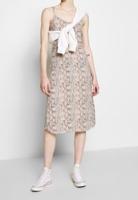 Midimekko, jossa on käärmeennahkapainatus beigen sävyissä, V-aukko ja ohuet olkaimet. Valkoinen collegepaita on sidottu rennosti hartioiden ympäri.