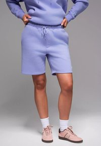 Shorts en sweat-shirt violet clair avec une taille élastique et un cordon de serrage. Associés à des chaussettes blanches et des baskets rose clair. Texture lisse.