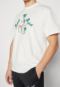 Camiseta de algodón blanca con un diseño pixelado verde y el logo de Nike en naranja en el centro. Cuello redondo y mangas cortas.