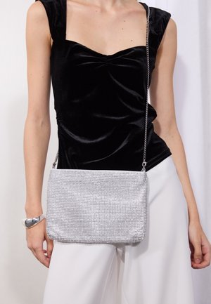 Personne portant un haut sans manches en velours noir, un pantalon blanc, un bracelet argenté, et tenant un sac bandoulière en chaîne argentée au niveau de la hanche.