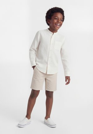 Smiješeći se dječak u bijeloj košulji s dugim rukavima na kopčanje, bež kratkim hlačama i bijelim slip-on cipelama stoji na jednobojnoj bijeloj pozadini.