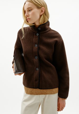 Un mannequin blond porte une veste en peau lainée marron foncé et tient une pochette en cuir marron sur un fond blanc uni.