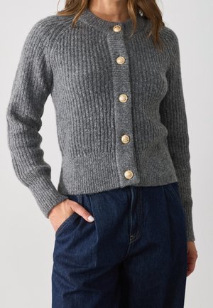 Femme portant un cardigan gris en tricot avec des boutons dorés et un pantalon bleu marine taille haute, main dans la poche.