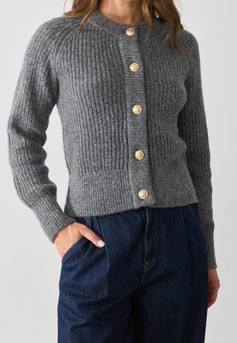 Femme portant un cardigan gris en tricot avec des boutons dorés et un pantalon bleu marine taille haute, main dans la poche.