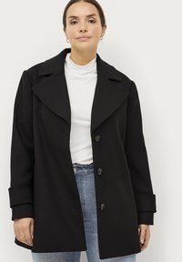 Zwarte, oversized jas met grote revers, vier knopen, zijzakken en een gladde textuur, gedragen over een witte coltrui en spijkerbroek.