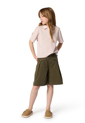 Jong meisje met lang haar, gekleed in een lichtroze shirt met korte mouwen, olijfgroene korte broek tot op de knie en beige instapschoenen, staand met de handen achter haar rug.