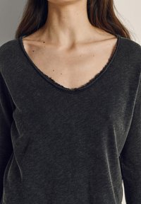 Nainen, joka pukeutunut tummanhiilenharmaaseen V-kaulaiseen pitkähihaiseen paitaan, jossa on raakareunainen kaula-aukko ja näkyviä ihon pisamia rinnassa ja kaulassa.