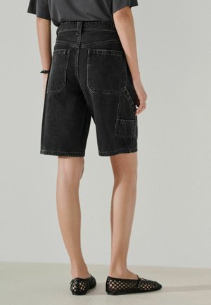 Person trägt schwarze, knielange Jeansshorts mit großen Taschen hinten und an den Seiten, schwarze flache Schuhe aus Mesh und ein dunkelgraues Oberteil, steht.