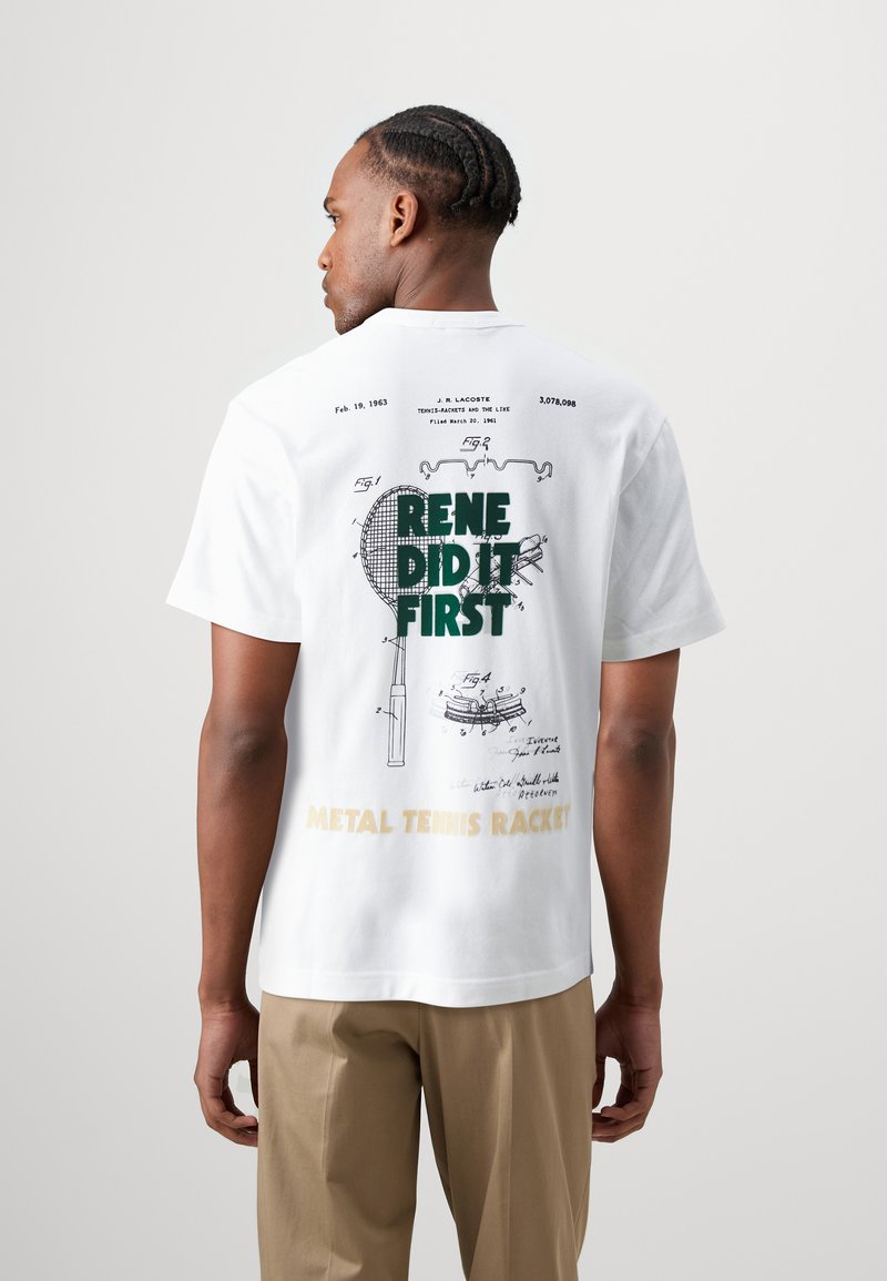 T-shirt in cotone bianco con stampa grafica di un brevetto per racchetta da tennis, testo verde "RENE HA FATTO PRIMA" e dettaglio dorato "RACCHETTA DA TENNIS IN METALLO".