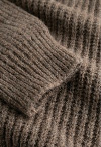 Brūns ribots adījums ar tuvplānu uz salocītu piedurknu uz mīkstas, teksturētas vilnas apģērba.