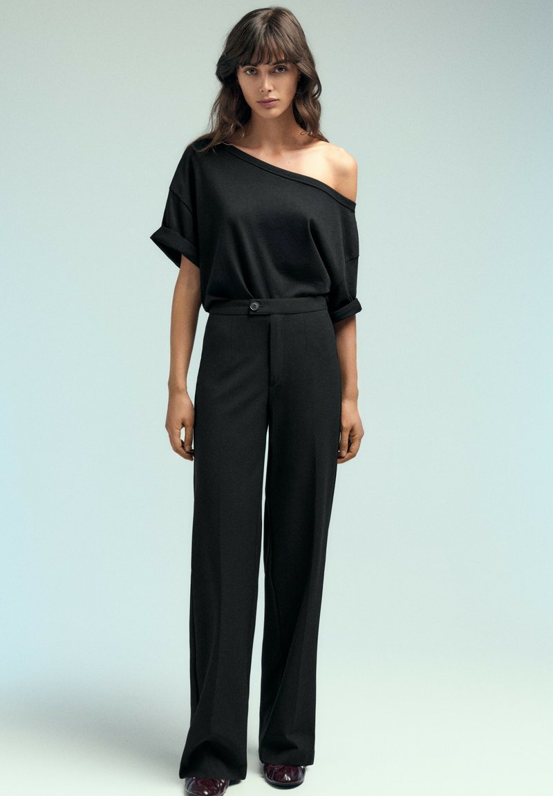 Femme debout portant un haut noir épaules dénudées, un pantalon noir large taille haute et des chaussures foncées sur un fond clair uni.
