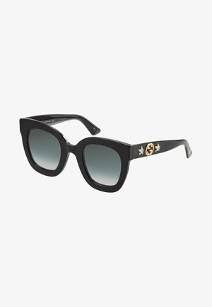Óculos de sol oversized pretos com uma armação grossa, lentes degradê e acessórios em dourado, apresentando um logótipo circular e detalhes de estrelas nas hastes.
