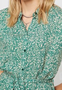 Camicia verde con motivo a fantasia e design maculato bianco, dotata di un bottone frontale, maniche corte e un laccio in vita per una forma aderente.