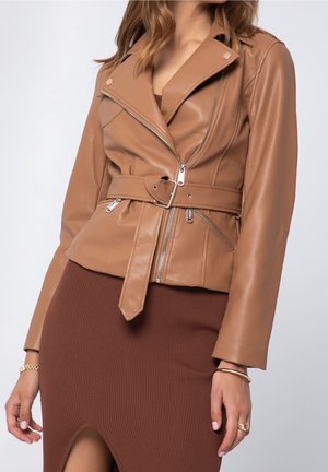 Frau in einer hellbraunen Lederjacke mit Gürtel und silbernen Reißverschlüssen über einem braunen gerippten Rock mit Schlitz vorne, goldenem Armband und Ringen.