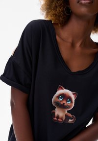 Zwarte T-shirt met korte mouwen, voorzien van een cartoonkat graphic met grote blauwe ogen en bruine accenten. Het shirt heeft een ontspannen pasvorm en een zachte textuur.