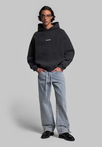 Felpa nera oversize con tasca frontale e logo bianco, abbinata a jeans azzurri a gamba larga e scarpe nere. Tessuto morbido, vestibilità rilassata.